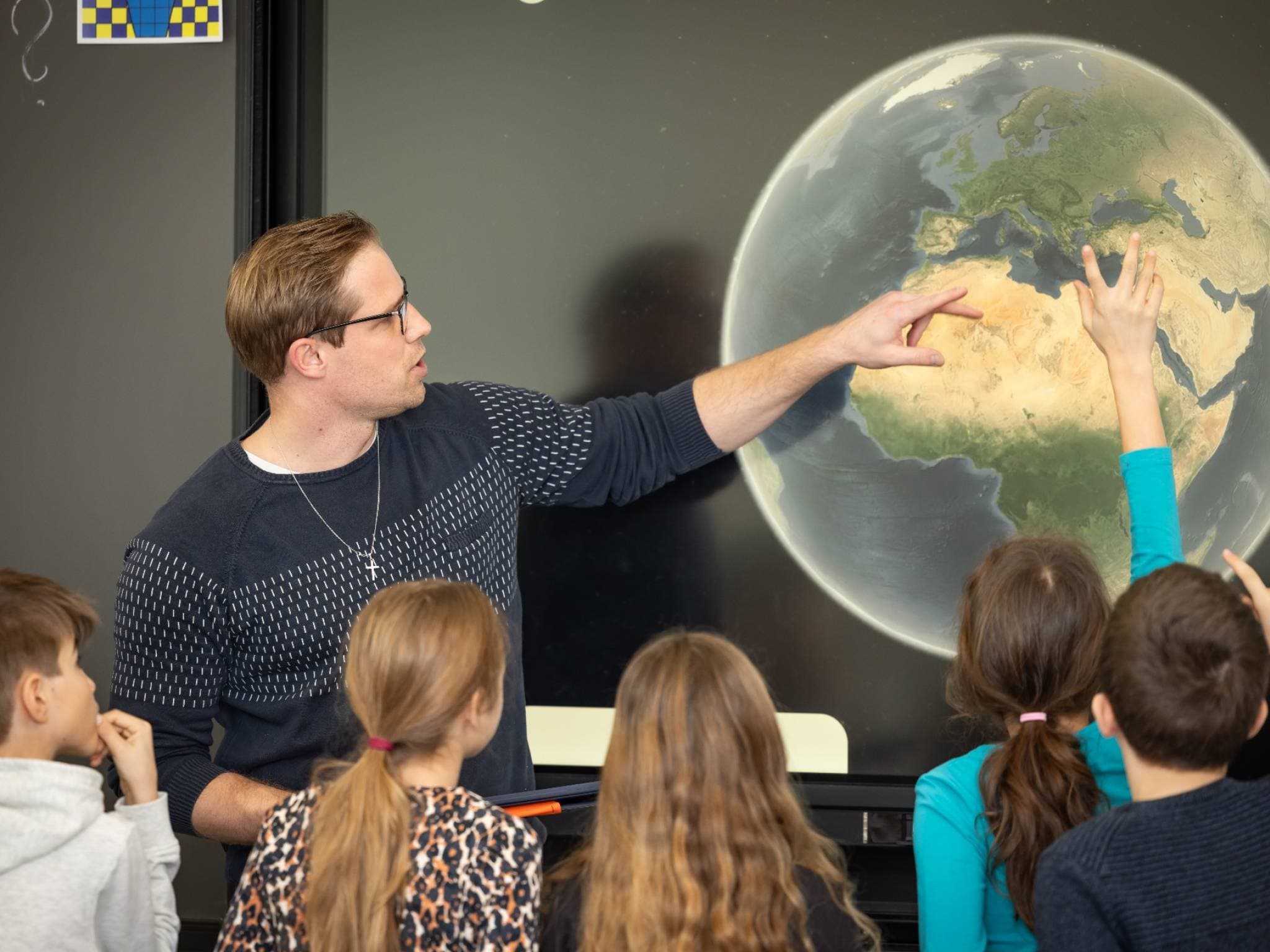 Samuel Schreiber im Unterricht: Er zeigt an der digitalen Wandtafel auf einen Globus, die Klasse hört zu, ein Kind meldet sich.